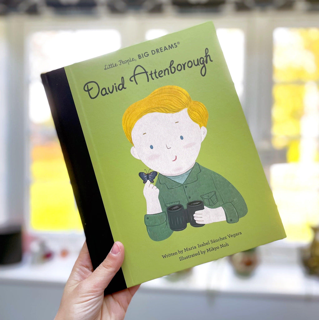 Picture of the book being held up at an angle by a hand. The book has a black spine. The front cover is a light green with "Little People, BIG DREAMS" and then David Attenborough's name underneath. There's an illustration of him. He is holding a black and blue butterfly in one hand and a pair of black binoculars in the other. He has a green button-up and a light green t-shirt underneath. At the bottom it credits the author Maria Isabel Sánchez Vegara. The Illustrator is Mikyo Noh. 